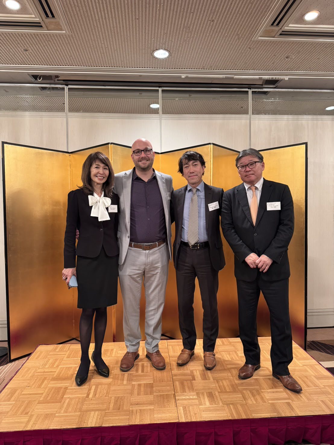 Four professionals pose together on a wooden stage, with a gold folding screen in the background. The group smiles, dressed in formal attire.
