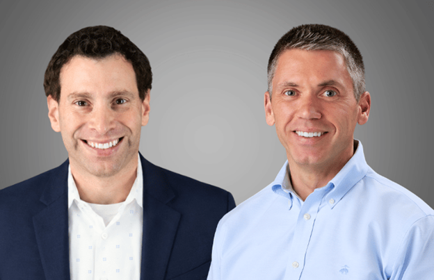 Two men smile at the camera, one in a dark blazer over a light shirt, the other in a light blue button-up shirt, with a grey gradient background.