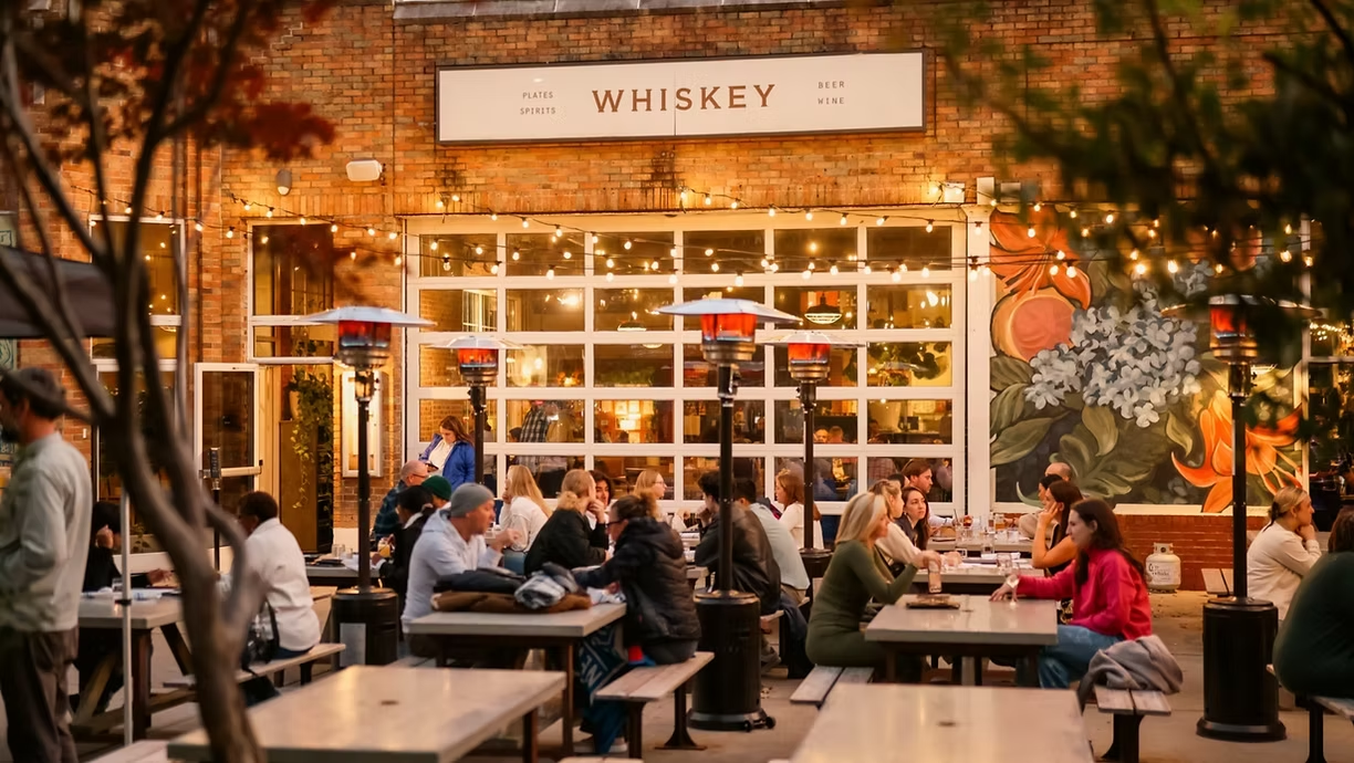 Outdoor dining at a venue named Whiskey, featuring string lights, patrons at wooden tables, and a floral mural on the wall.