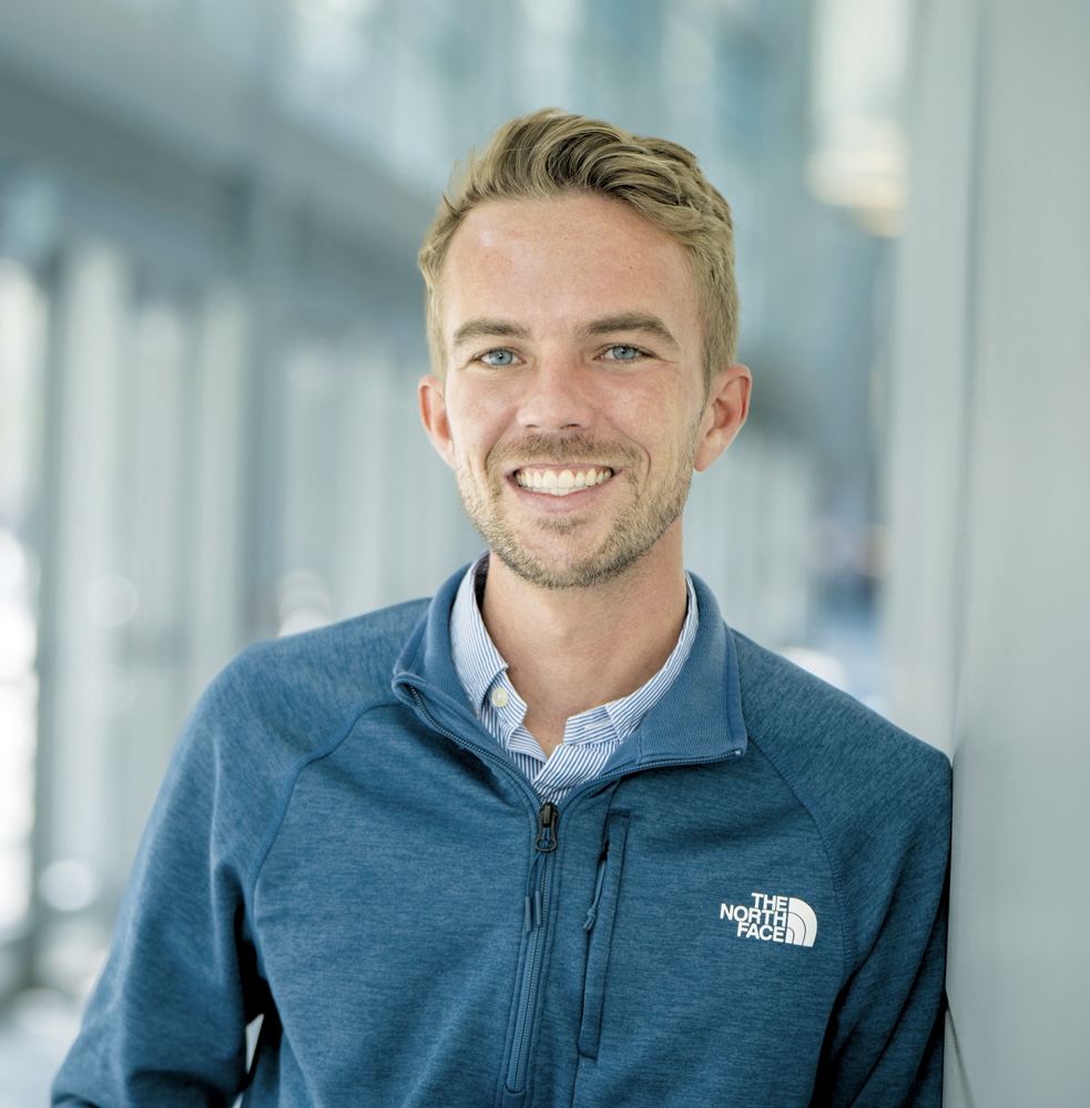 Smiling young man in a blue North Face jacket stands against a light grey background, with natural light highlighting his cheerful expression.